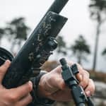 Närbild på en jägare som monterar Spartan Javelin Pro Hunt Tac benstöd i ett magnetiskt snabbfäste på ett gevär ute i naturen.