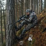 En jägare i kamouflagekläder sitter i en brant skogssluttning och siktar med ett gevär monterat på ett Spartan Springbok bipod benstöd.