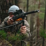 En jägare som siktar med gevär monterat på ett Spartan Springbok bipod i en skogsmiljö sett framifrån.
