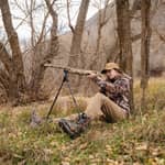 En jägare i kamouflagekläder sitter på marken i en skog och siktar med ett gevär monterat på ett Spartan Springbok bipod-stöd.
