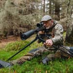 En jägare i kamouflagekläder sitter på skogsmark och siktar med ett gevär monterat på ett Spartan Springbok Quad skjutstöd.