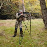 En jägare i kamouflagekläder siktar med ett gevär monterat på ett Spartan Springbok Quad skjutstöd i en skogsmiljö.