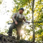 En jägare håller i en hopfälld Spartan Springbok Tripod i en skogsmiljö vid solnedgång.