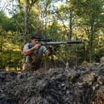 En jägare som siktar med ett gevär monterat på ett Spartan Springbok tripod-stativ i en skogsmiljö.