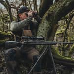 En jägare som sitter i skogen och spanar med kikare medan geväret vilar stabilt på ett Spartan Springbok stativ.
