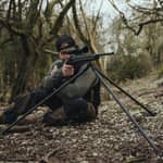En jägare i sittande skjutställning med ett gevär monterat på ett Spartan Springbok Tripod kolfiberstativ i skogsterräng.