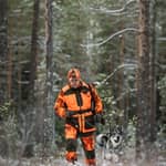 Jägare i orange Pinewood Furudal Doghandler-väst med kamouflagemönster och matchande keps går genom en snöpudrad tallskog med kopplad hund i grönt koppel