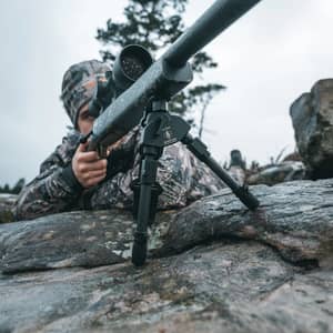 En jägare i kamouflagekläder siktar med ett gevär monterat på ett Spartan Javelin Pro Hunt Tac benstöd på en stenhäll.