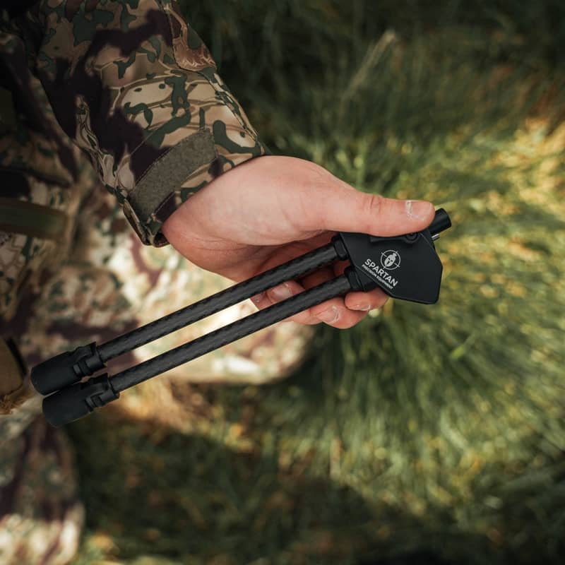En jägare håller ett Spartan Javelin Lite benstöd i kolfiber i handen i en naturlig utomhusmiljö.
