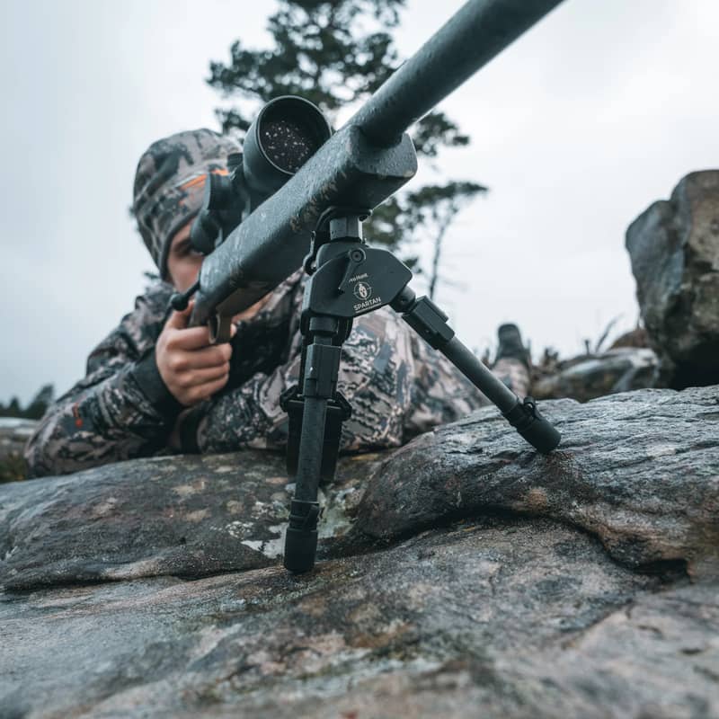 En jägare i kamouflagekläder siktar med ett gevär monterat på ett Spartan Javelin Pro Hunt Tac benstöd på en stenhäll.