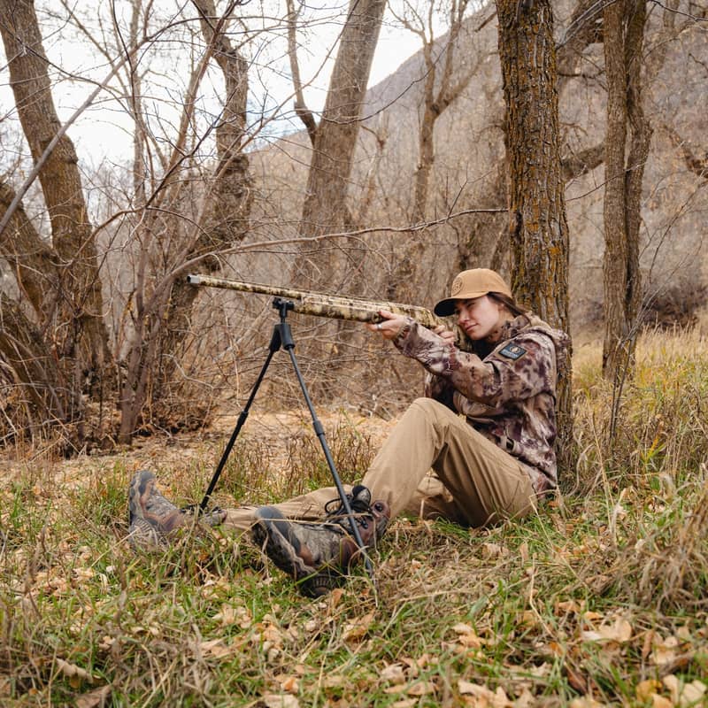 En jägare i kamouflagekläder sitter på marken i en skog och siktar med ett gevär monterat på ett Spartan Springbok bipod-stöd.