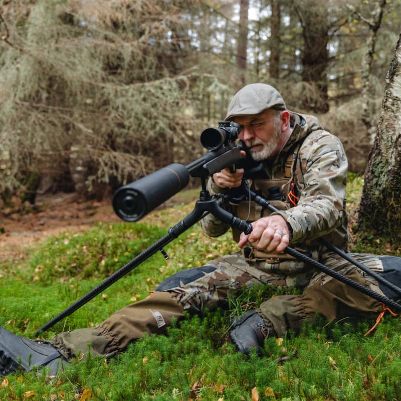 En jägare i kamouflagekläder sitter på skogsmark och siktar med ett gevär monterat på ett Spartan Springbok Quad skjutstöd.