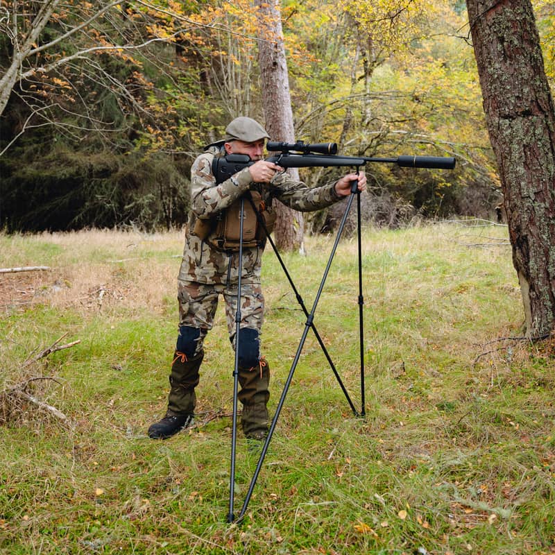 En jägare i kamouflagekläder siktar med ett gevär monterat på ett Spartan Springbok Quad skjutstöd i en skogsmiljö.