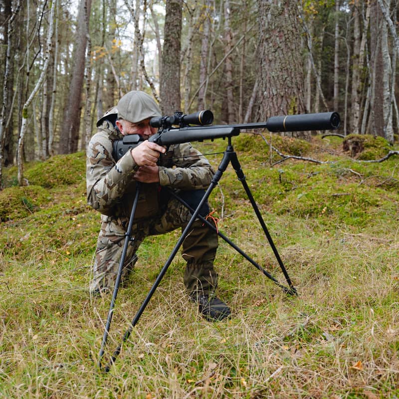 En jägare i kamouflagekläder siktar med sitt gevär monterat på ett Spartan Springbok Quad skjutstöd i en skogsmiljö.