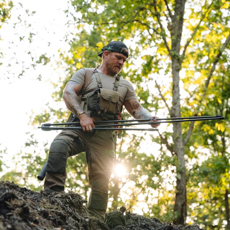 En jägare håller i en hopfälld Spartan Springbok Tripod i en skogsmiljö vid solnedgång.