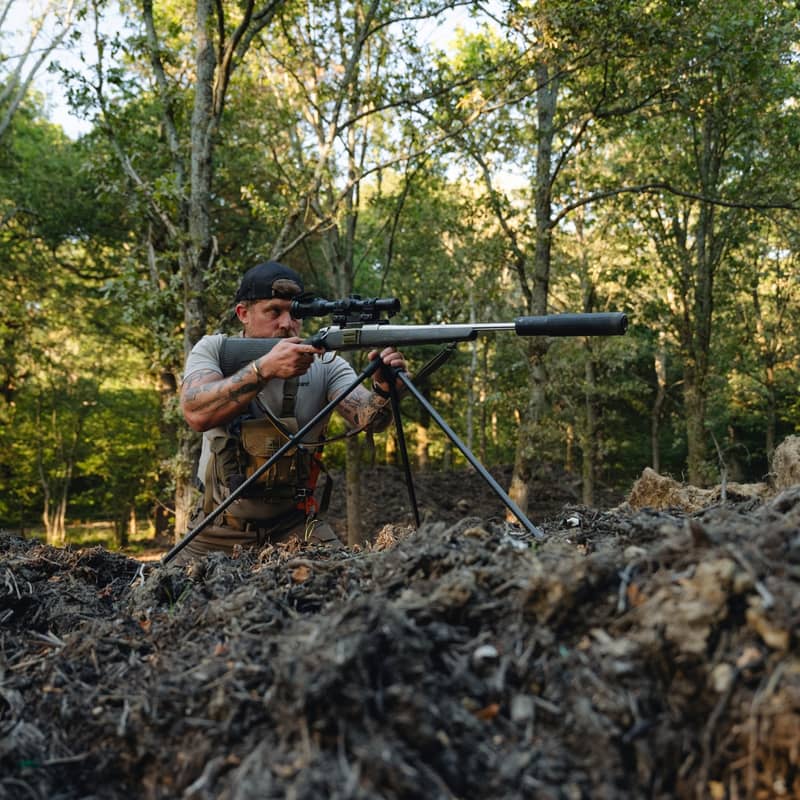 En jägare som siktar med ett gevär monterat på ett Spartan Springbok tripod-stativ i en skogsmiljö.