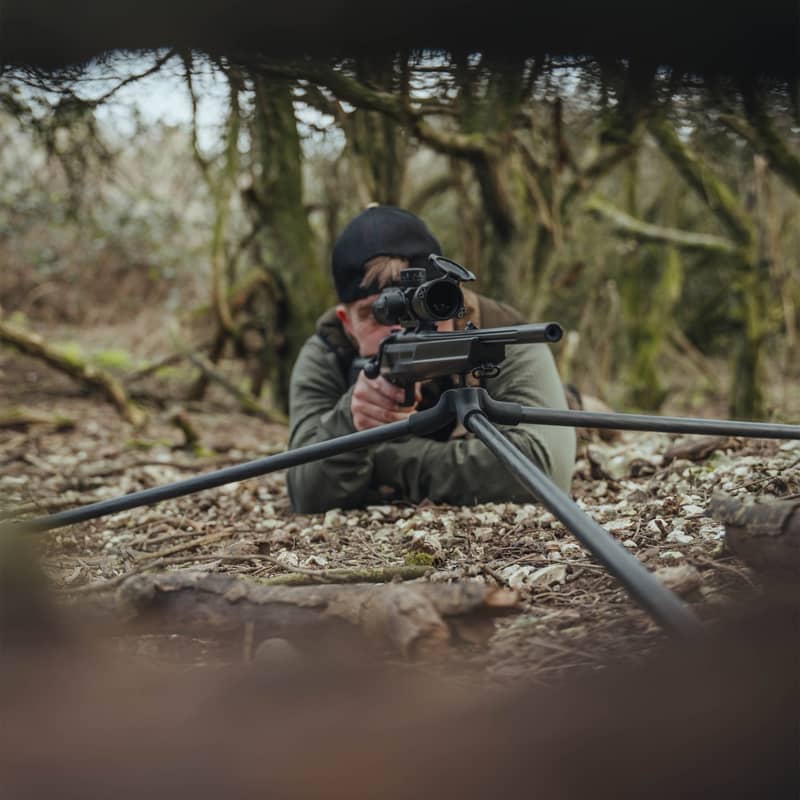 En jägare som siktar med sitt gevär monterat på ett Spartan Springbok tripodstativ i liggande position i skogen.