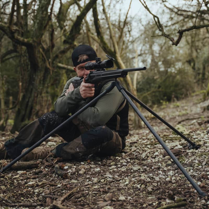 En jägare i sittande skjutställning med ett gevär monterat på ett Spartan Springbok Tripod kolfiberstativ i skogsterräng.