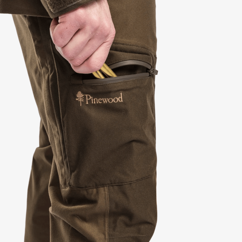 Närbild på benficka med dragkedja och broderad Pinewood-logga på bruna Pinewood Furudal Active 2L byxor, med hand som drar fram patroner ur fickan