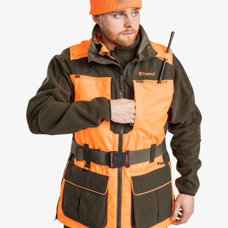 Detaljbild av Pinewood Furudal Doghandler väst i orange och grön färg, buren av modell, med radioantenn i bröstficka och justerbart midjebälte.