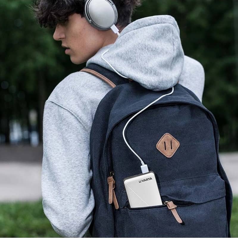 Ung person med grå huvtröja och hörlurar bär en marinblå ryggsäck med en vit Varta powerbank i framfickan, kopplad med en vit laddkabel till hörlurarna