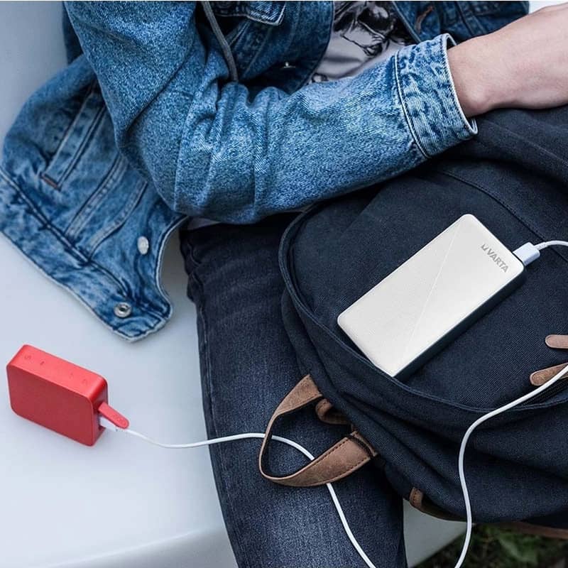 Vit Varta powerbank som ligger på en mörkblå ryggsäck i en persons knä och laddar en röd bärbar högtalare med en vit USB-kabel