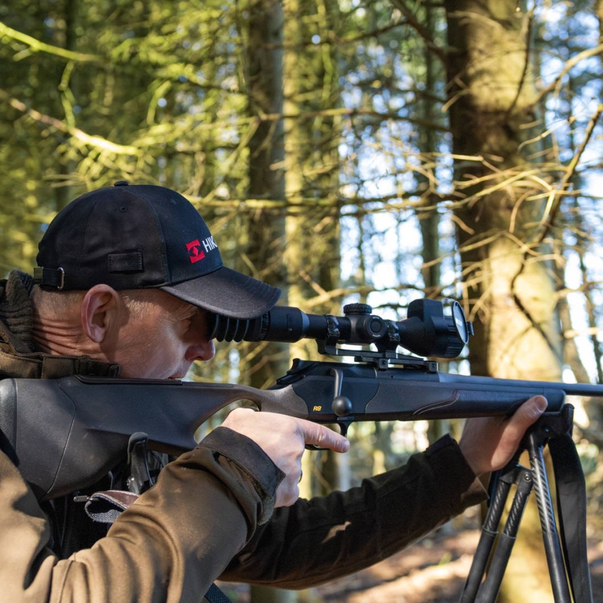 Hikmicro NEOS NH25L monterat på gevär i skogsmiljö Jägare i keps sikter genom ett Hikmicro NEOS NH25L termiskt riktkikare monterat på ett gevär med stödben i solbelyst skog