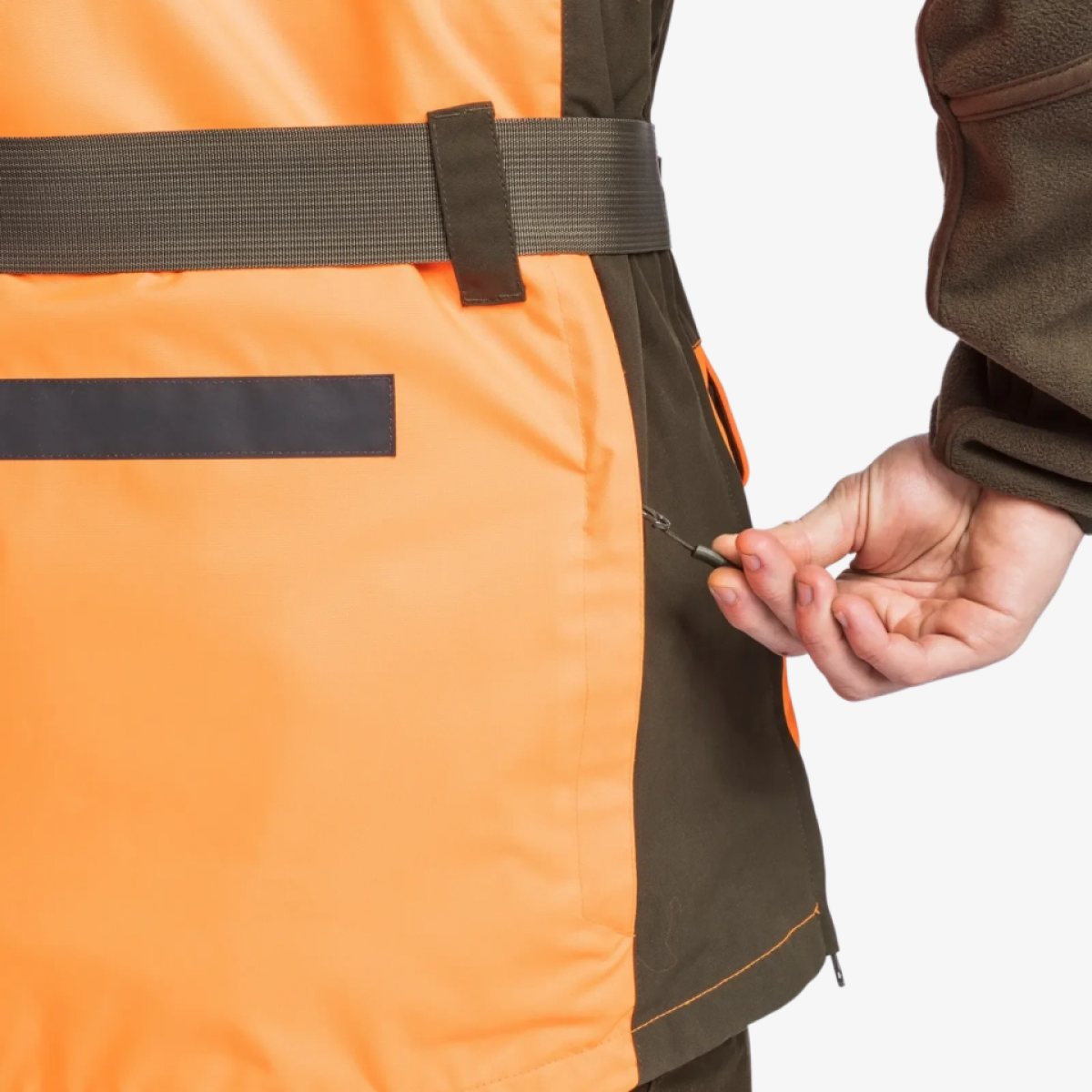 Furudal Doghandler väst – sidodetalj med dragkedja Närbild på sidan av Pinewood Furudal Doghandler väst i orange och mörkgrönt med midjerem, reflexband och en hand som drar i dragkedjan vid sidan